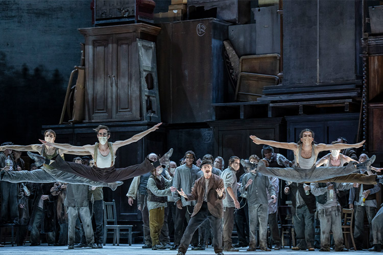 The company of Fiddler on the Roof. Two dancers are in the front performing split leaps.